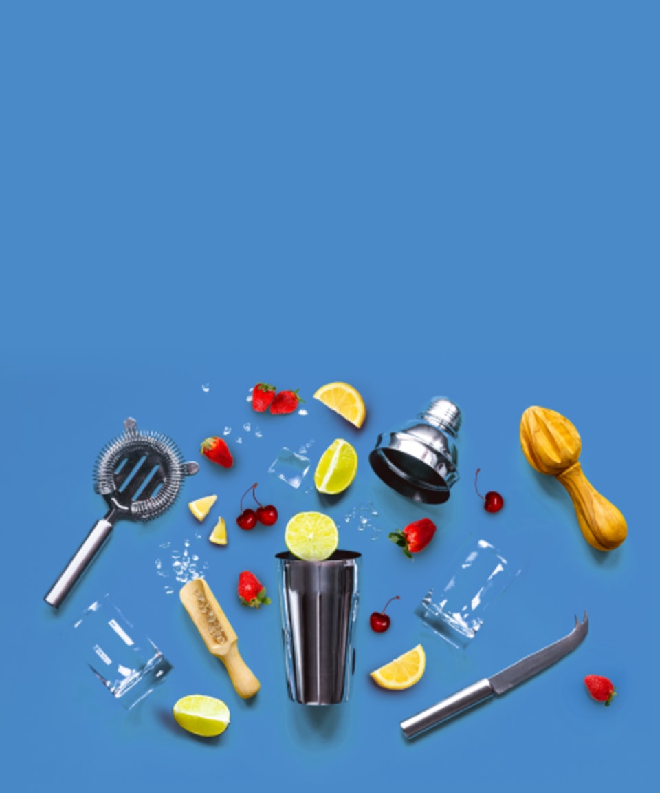 A cocktail shaker surrounded by various fruits, citrus reamers, glasses, ice, and a knife for Confetti's Virtual Mixology Classes