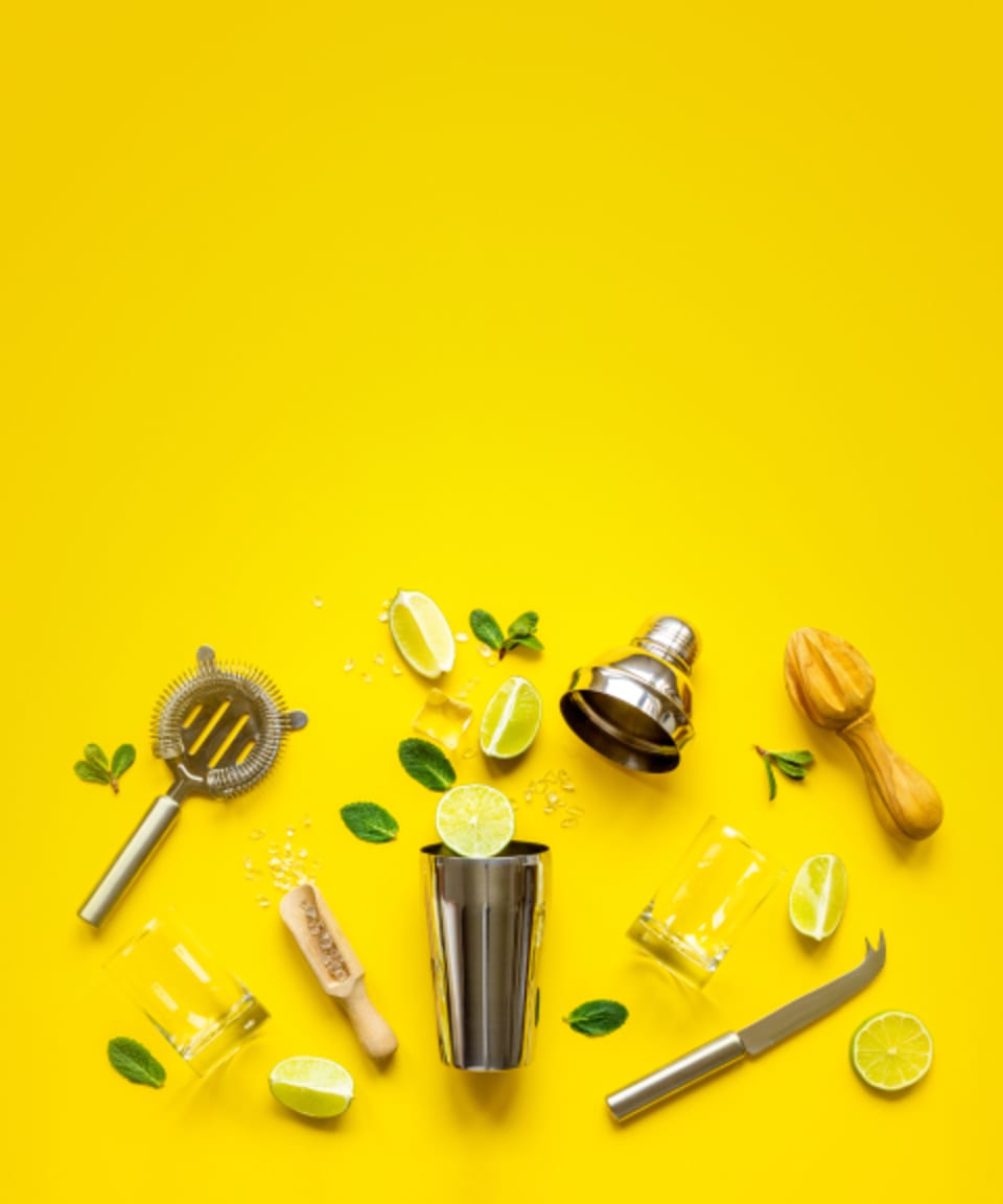 A cocktail shaker, strainer, knife, ice, a rocks glass, a scooper, sprigs of herbs, and slices of limes, all spread out for Confetti's BYO Mixology Class