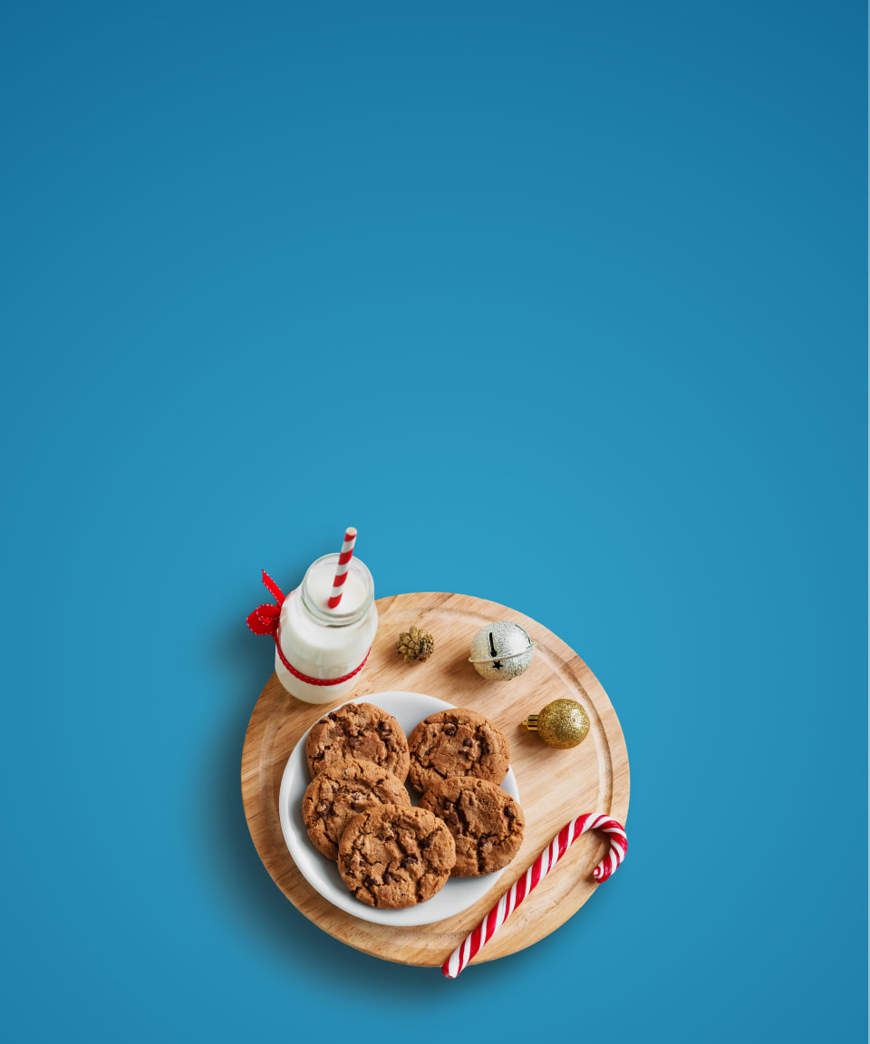 A plate of chocolate chip cookies, a candy cane, and a glass bottle of milk sitting on a wood cutting board with ornaments for Confetti's Virtual BYO Holiday Baking Class