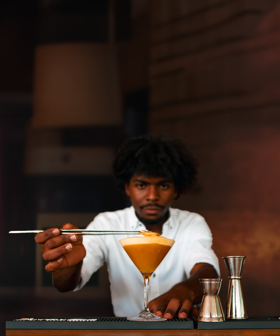 Black mixologist using tongs to place garnishes on a cocktail next to a shaker and jigger 