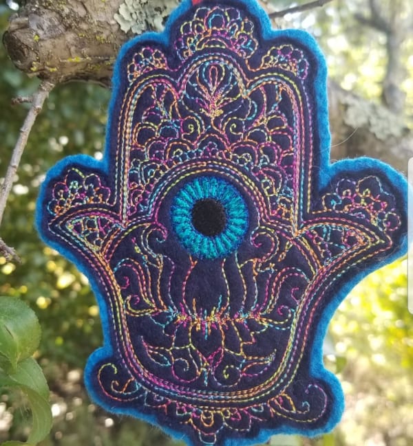 This Hamsa is a palm-shaped good luck talisman for your door or wall. It is a symbol of protection, to ward off the "evil eye", and bring good luck!
I hand-digitized the embroidered design on navy blue felt, using multi-color thread. The back is lined with a turquoise wool felt.
It hangs from a red suede loop, and is embellished with a silver-tone hamsa bead, and glass beads. 