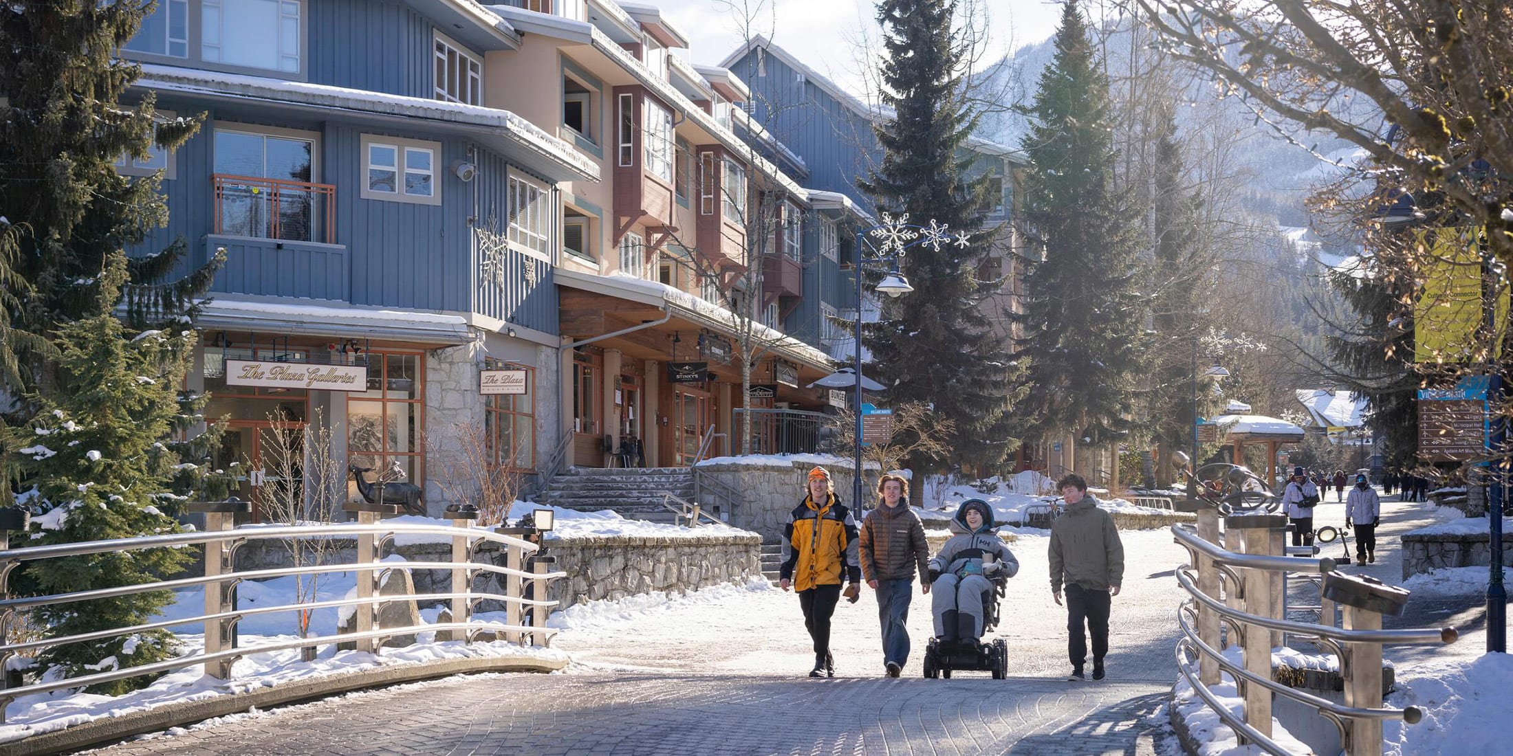 A group of friends, including one in a wheelchair, enjoys a sunny winter day in Whistler Village, with snow-covered mountains and cozy accommodations in the background