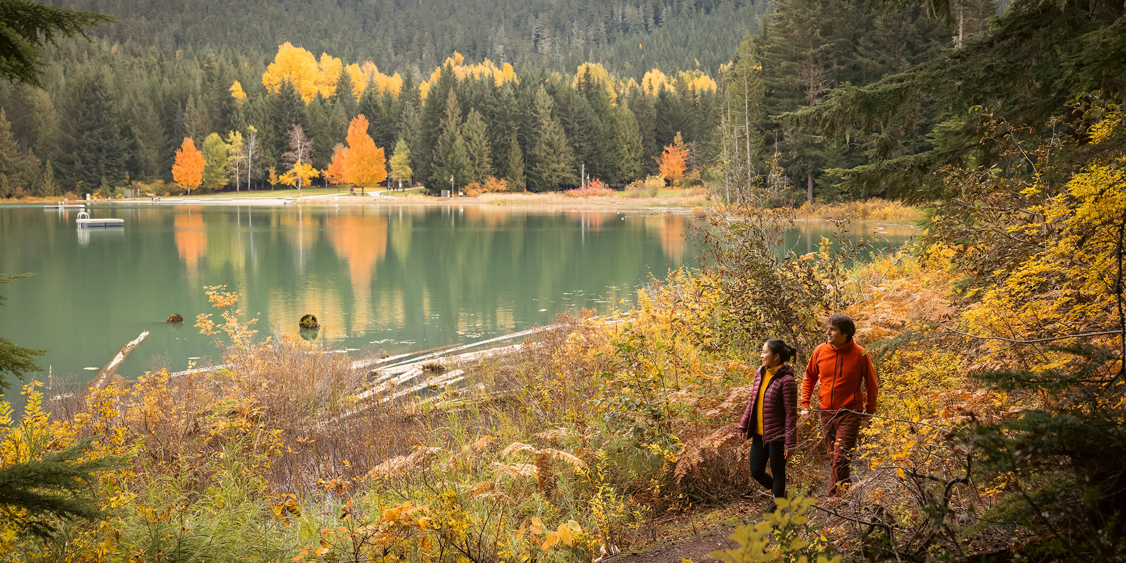 Two hikers strolling a forested lakeshore trail in autumn in Whistler, BC, surrounded by golden fall foliage and a reflective mountain lake.