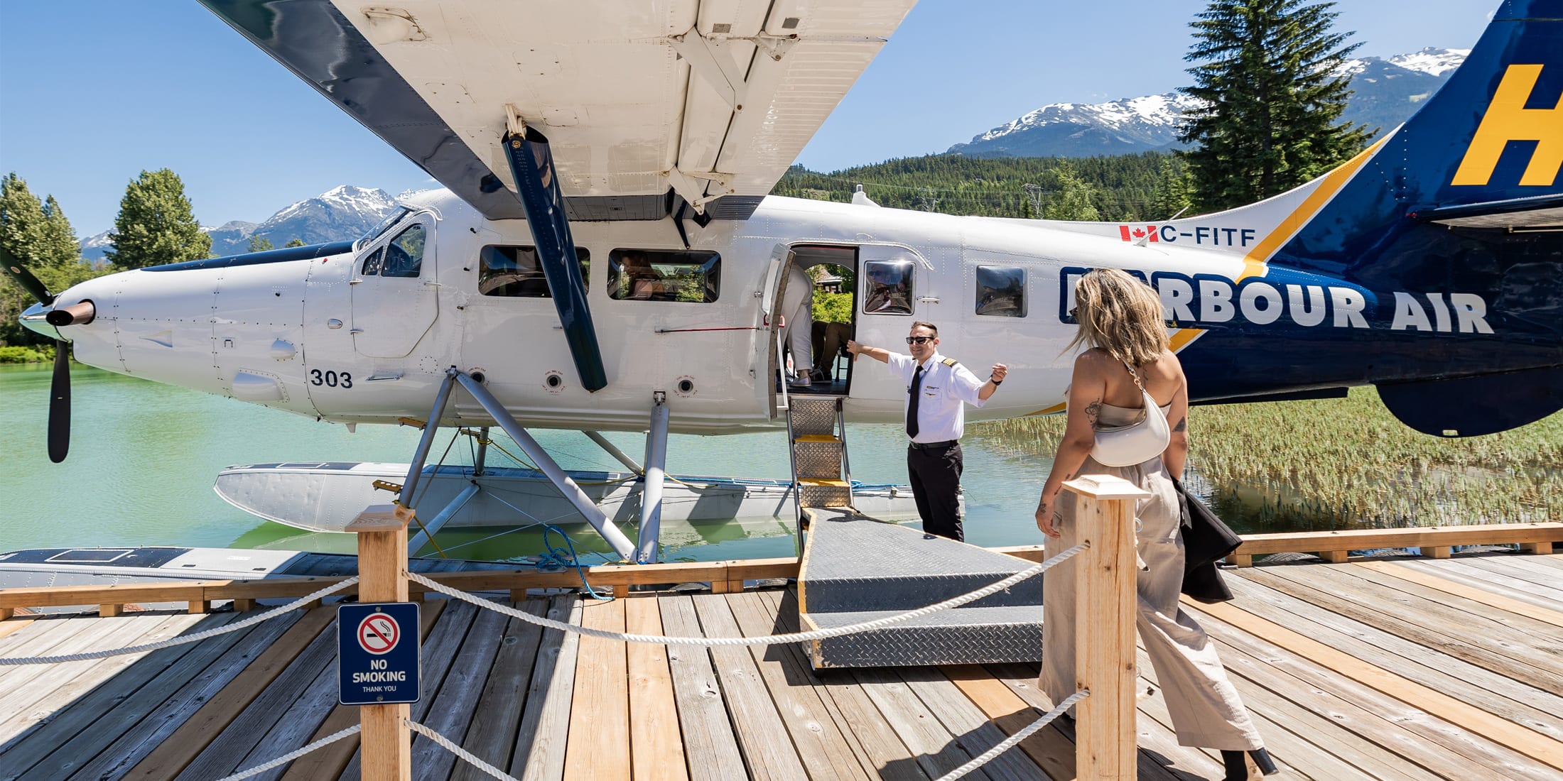 A Harbour Air floatplane docked on a lake in Whistler as a pilot welcomes a guest boarding via a small ramp, with turquoise water, forested shoreline and snow-capped Coast Mountains in the background.
