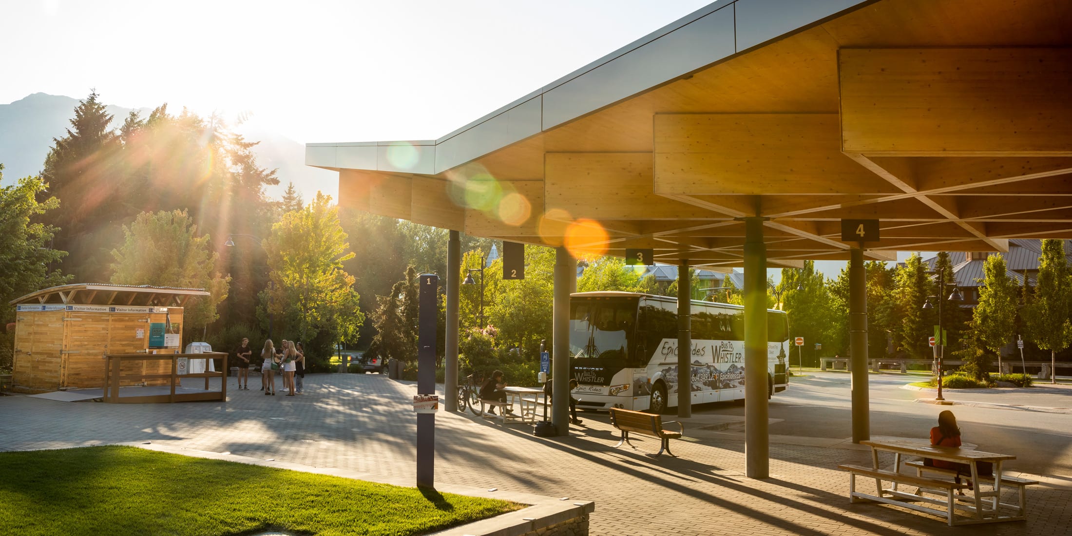 Bus loop in Whistler Village at golden hour, with a tour bus parked under a modern wooden canopy, numbered pickup bays, and people waiting or relaxing nearby in warm evening sunlight filtering through trees.