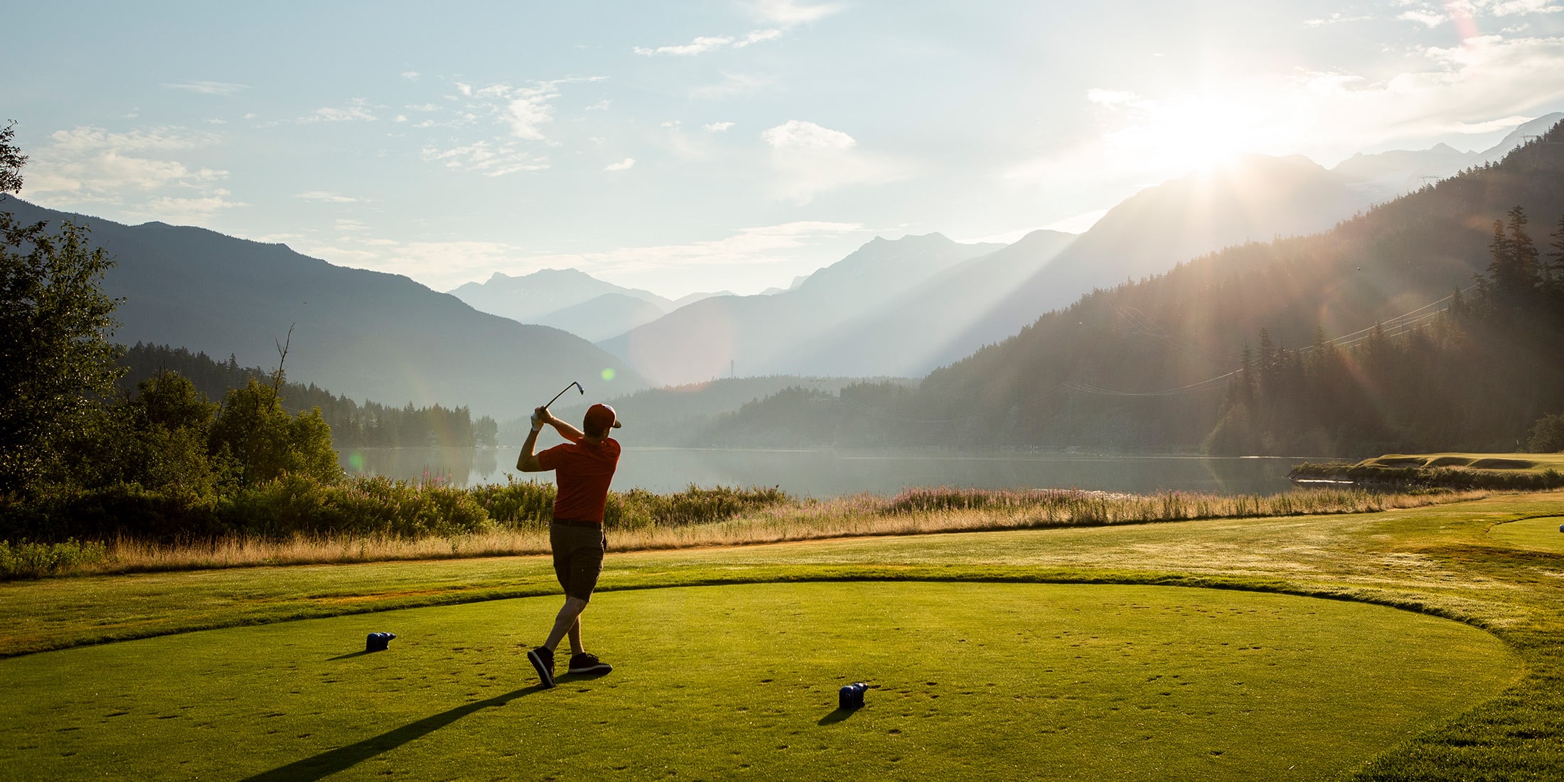A golfer tees off on a lush green fairway in Whistler, surrounded by towering evergreens and majestic mountain views.