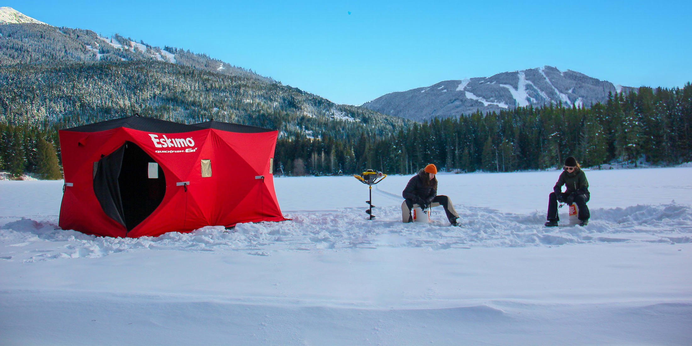 Two anglers ice fishing on buckets beside a red ice fishing tent and an auger on a snowy frozen lake, with forested ski mountains under a clear blue sky in Whistler.