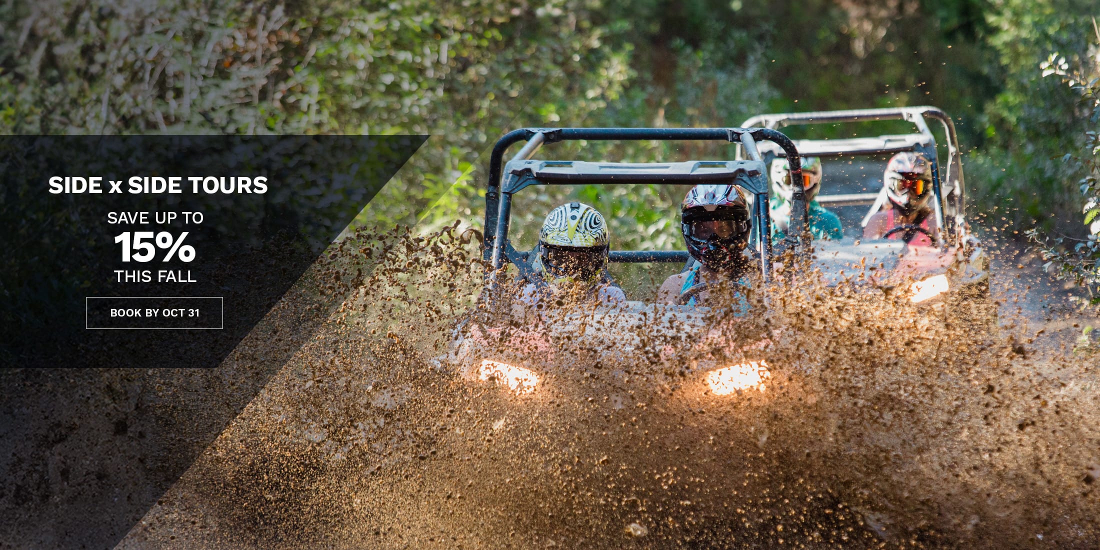 Two people ride in a side-by-side off-road vehicle splashing through mud on a forest trail, with additional riders behind.