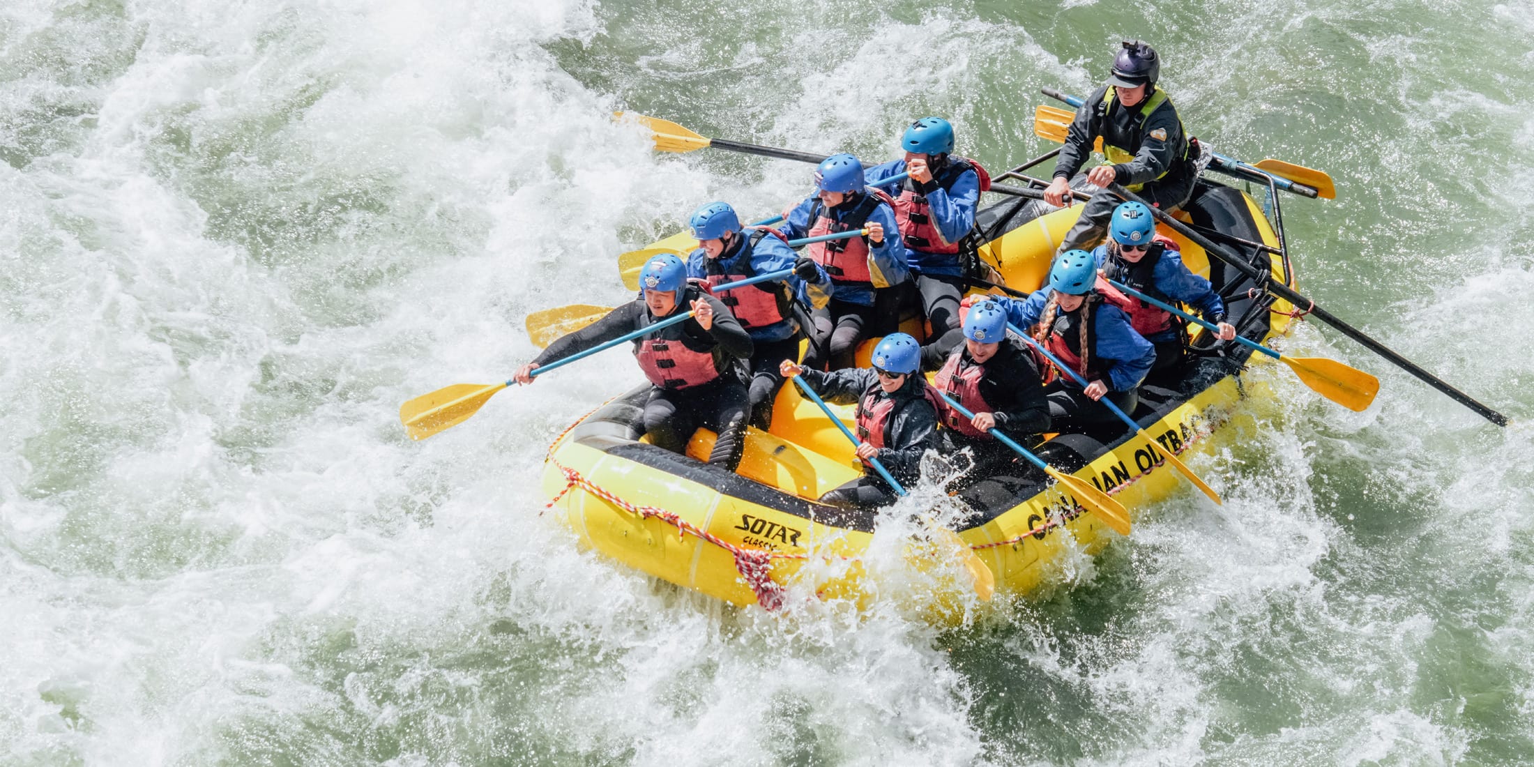 Group of rafters in orange life jackets paddle a red raft through fast-moving whitewater rapids.