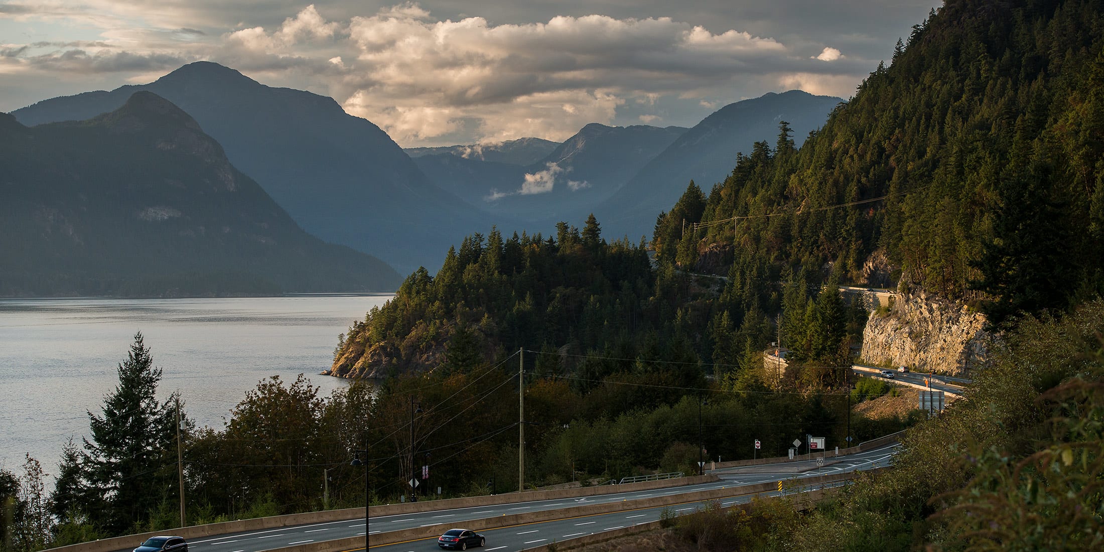 Scenic view of the Sea to Sky Highway winding along the coastline near Whistler, with forested mountains and Howe Sound at sunset.