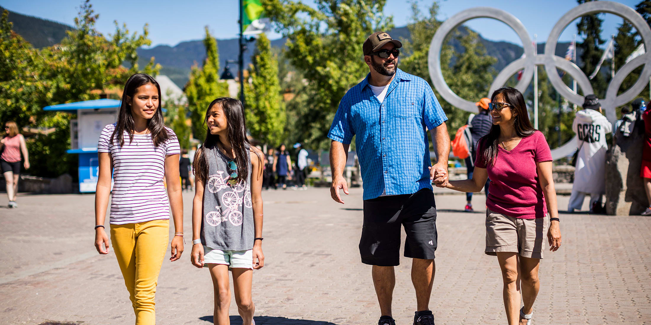 Family of four walking through Whistler Village on a sunny day, with mountains, trees and the Olympic rings sculpture in the background.