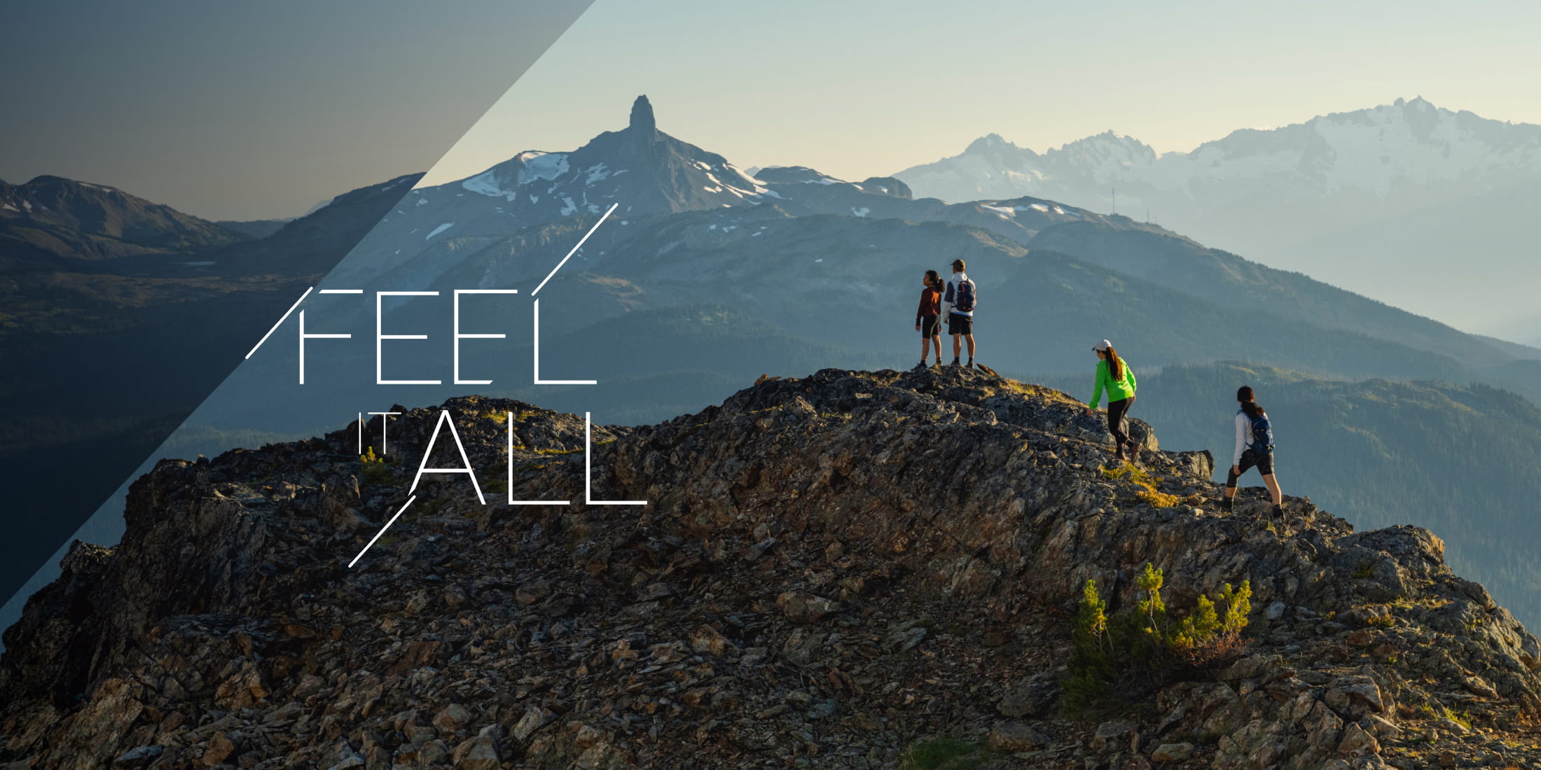 A group of hikers at the peak of Whistler Mountain with Black Tusk in the distance. 'Feel It All' overlay captures the essence of adventure in the mountains.