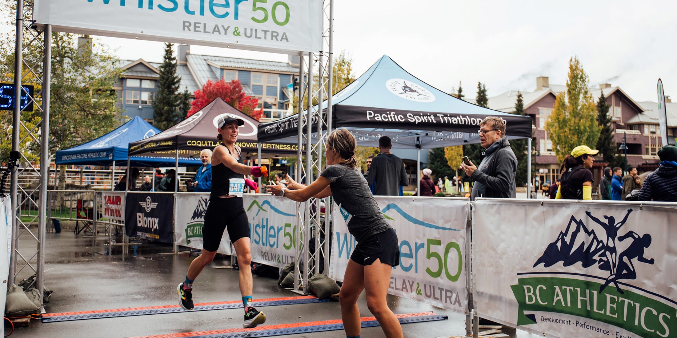 Runners exchanging a baton at the Whistler 50 Relay and Ultra finish line, surrounded by race banners, tents, and cheering spectators.