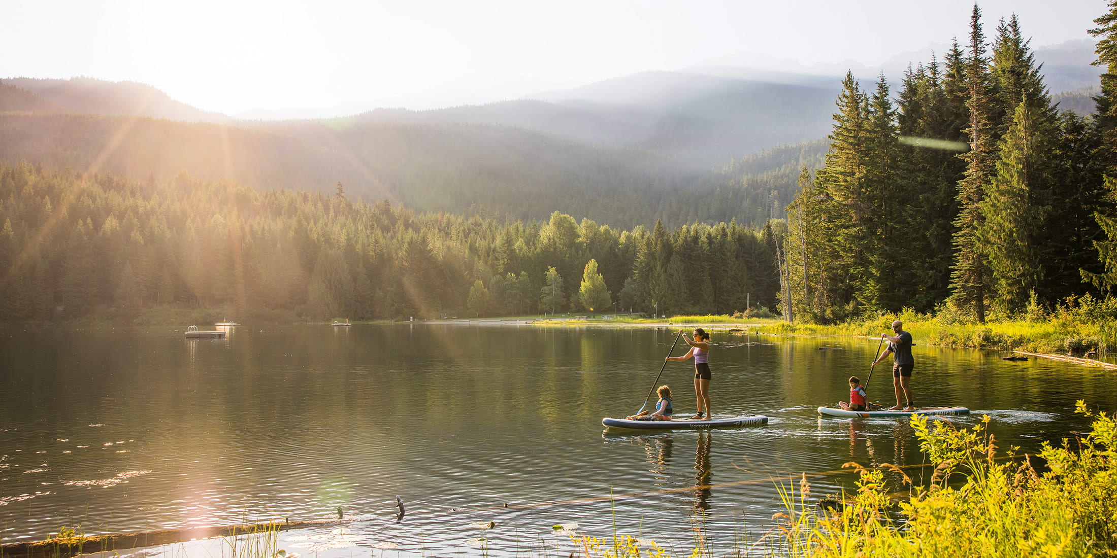 Two adults and two children paddleboarding on a calm lake in Whistler, surrounded by forest and mountains in warm evening sunlight.