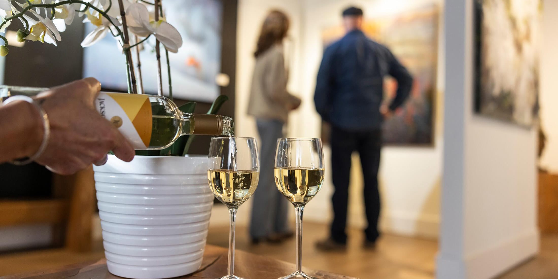 A couple perusing fine art at a gallery while someone pours white wine into a couple of glasses in Whistler BC Canada