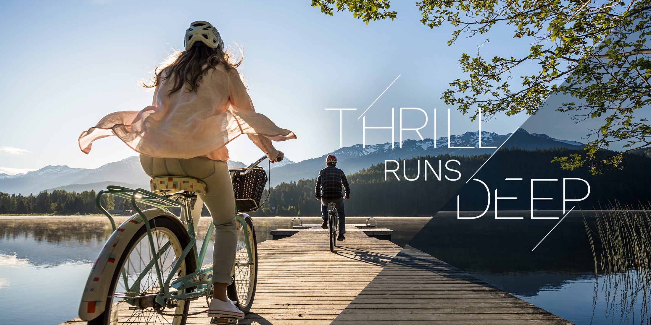 Two cyclists ride along a wooden dock at sunrise, framed by calm lake water and snow-capped peaks. Overlaid text reads ‘Thrill Runs Deep.