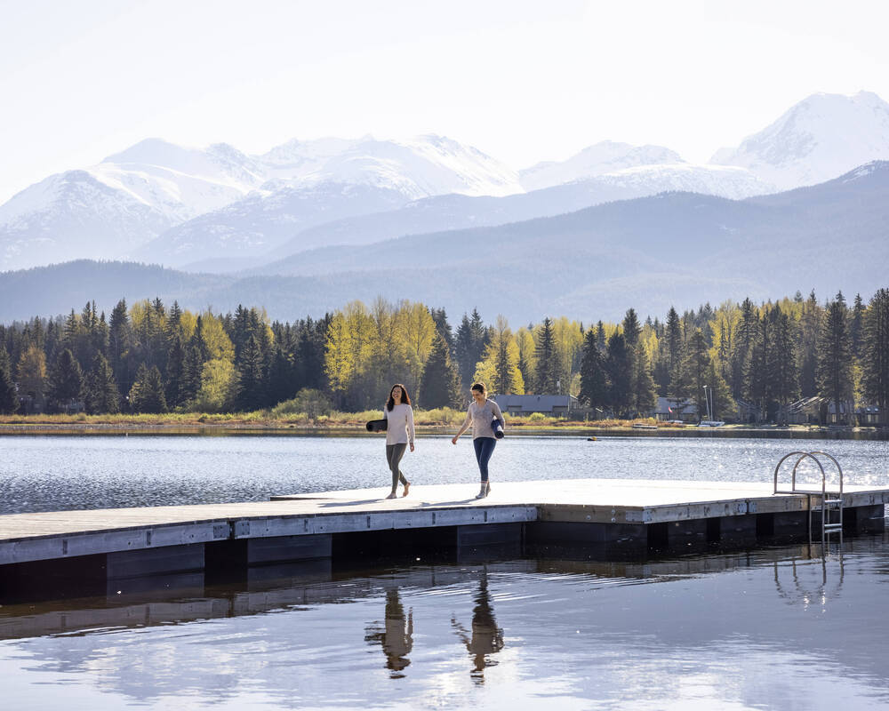 Two people walking on a lake dock with yoga mats in hand and mountains in the backgound