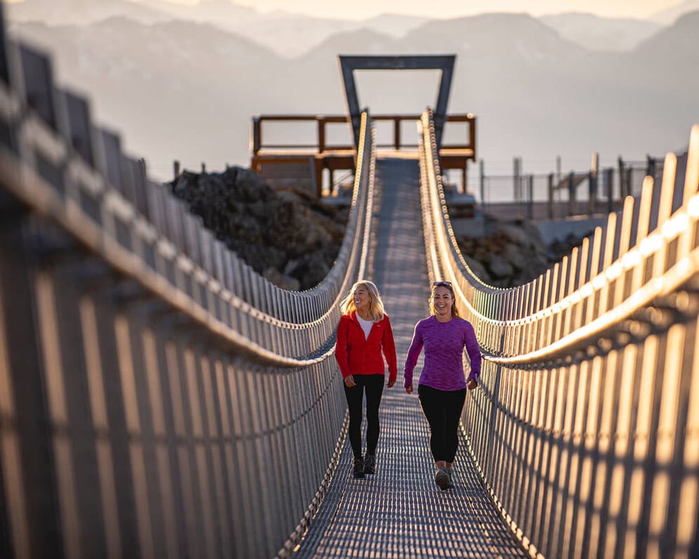 Two people walking across the Cloudraker Skybridge at the top of Whistler Mountain in summertime