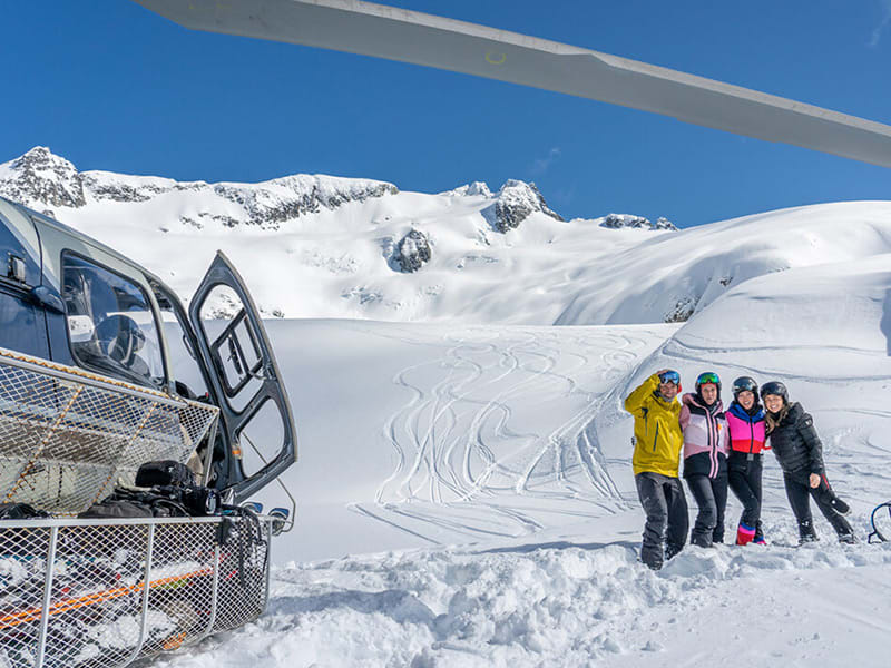 Group of skiers pose beside a Phantom Heli-Skiing helicopter on snowy alpine slopes near Whistler Blackcomb.