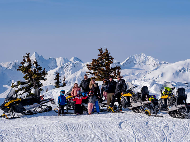 Group standing beside snowmobiles on a sunny Whistler ridge with panoramic snowy mountain peaks.
