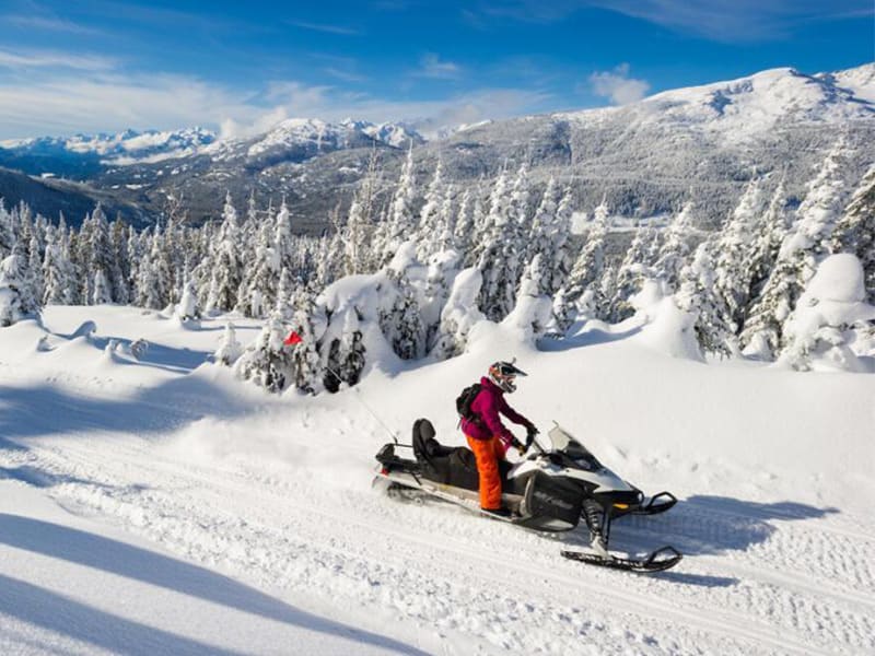Snowmobiler riding a sunny alpine trail overlooking snowy Whistler mountains and forest.