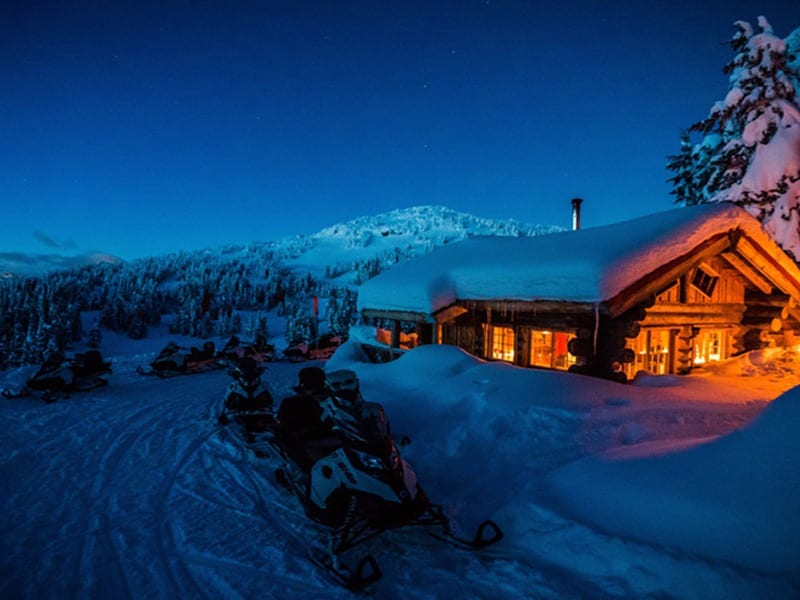 Snowmobiles parked outside a warmly lit Whistler log cabin under a deep winter night sky.