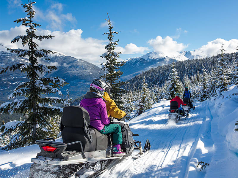 Snowmobilers riding a snowy Whistler trail with bright sunshine and panoramic mountain views.