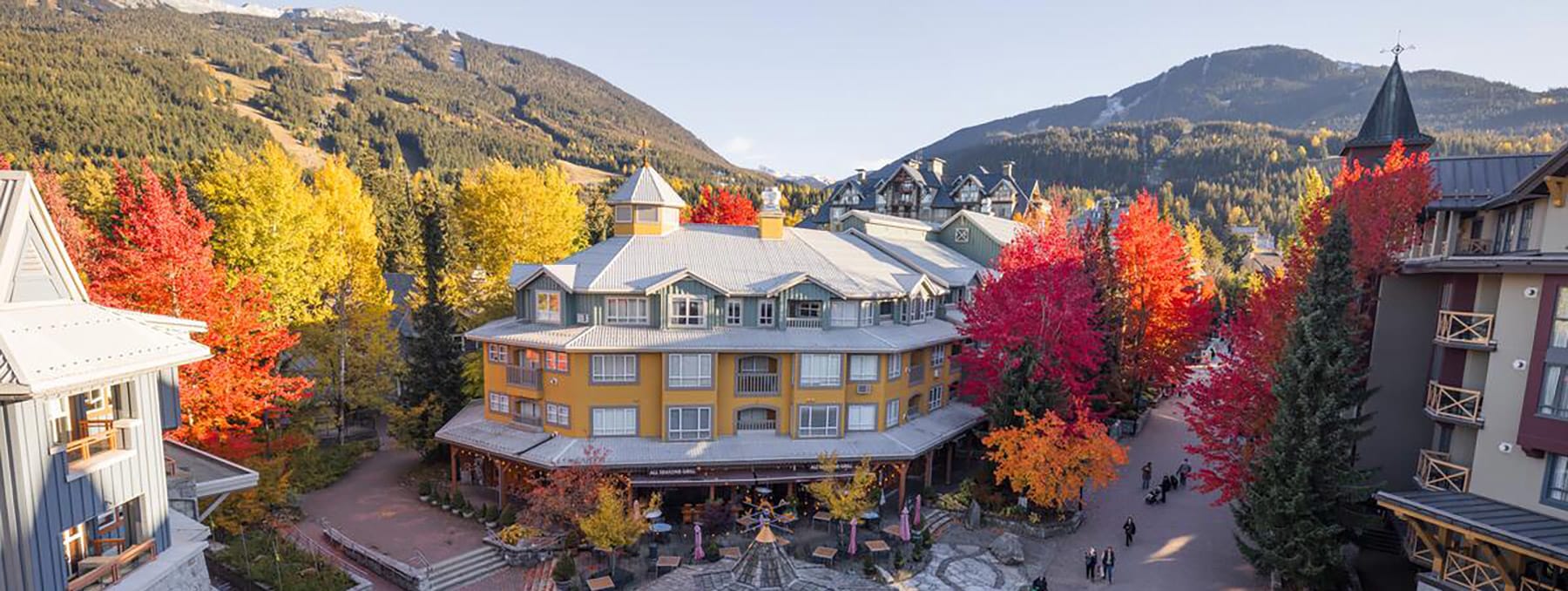 A view of Whistler Town Plaza in the fall with red and gold trees and Whistler Blackcomb in the background.
