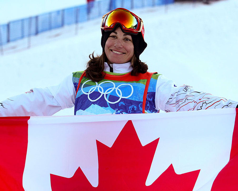 Winter sports athlete wearing an Olympic competition bib and ski goggles holding a Canadian flag on a snowy slope.