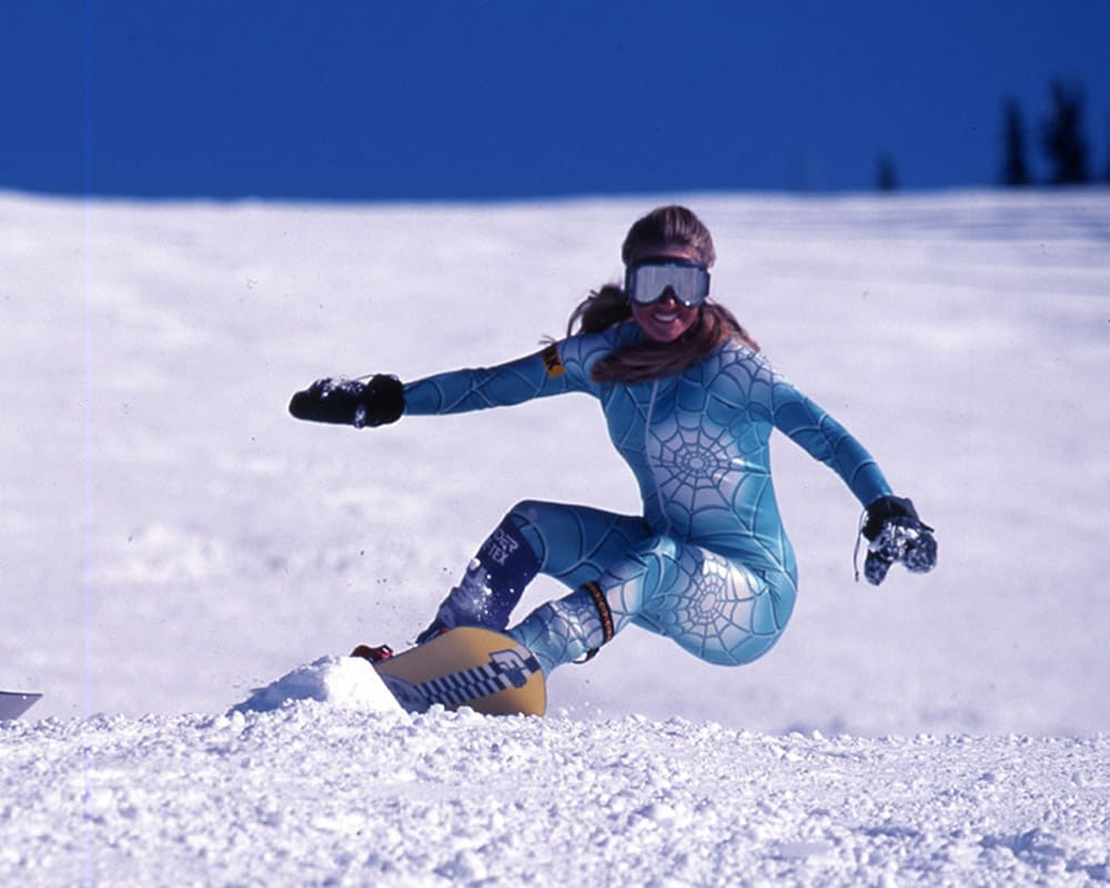 A snowboarder in a teal, spider-web–patterned race suit carving down a snow-covered Whistler, BC slope under a clear blue sky.