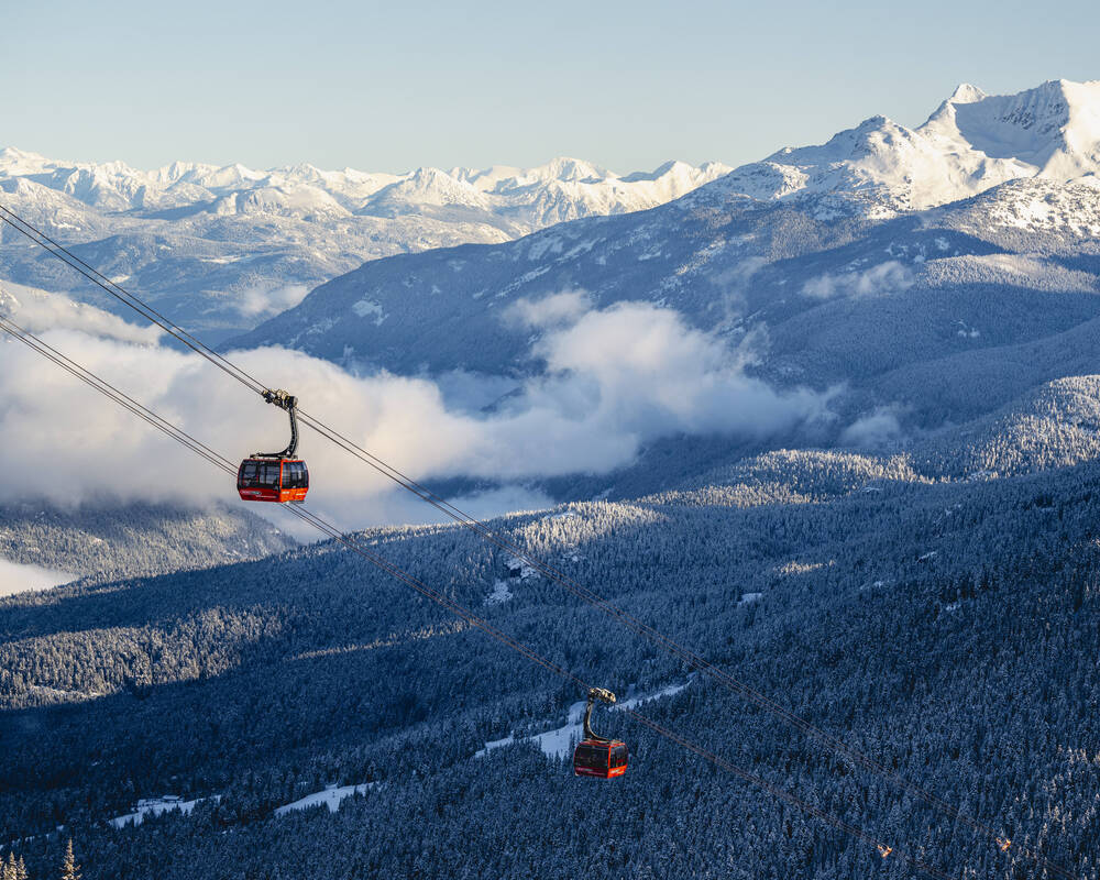 Two red Whistler Blackcomb Peak 2 Peak gondola cabins suspended over a snow-covered Coast Mountain forest in Whistler, BC, with panoramic snowy peaks in the background.