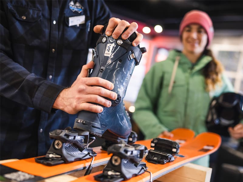 Ski rental technician fits a ski boot to bindings on orange skis while a skier waits in a Whistler rental shop.