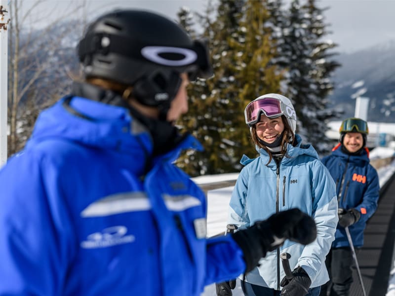 Ski instructor in a blue jacket speaking with three skiers on a sunny, snow-covered alpine slope, with panoramic mountain views in the background.