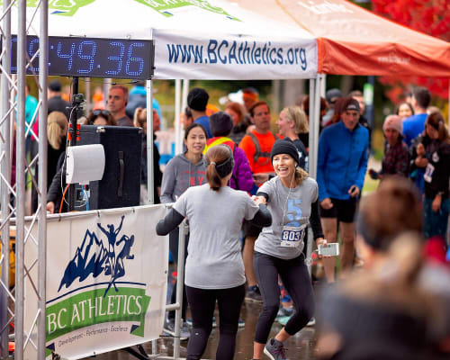 Runner crossing the finish line under a BC Athletics canopy with a digital clock reading 49:36, high-fiving a fellow participant amid cheering spectators in Whistler, BC.