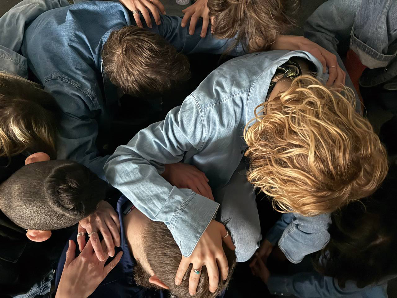 Overhead view of approximately eight people gathered in a tight huddle, heads bowed toward the center, bodies overlapping. A person with curly blonde hair wearing a light blue denim shirt is visible in the foreground, arm extended across another person's back. Several hands are visible resting on shoulders and backs. The group is dressed in casual denim clothing. The image is shot in warm natural light. The faces are hidden, the focus entirely on the physical contact between bodies in a moment of collective stillness, vulnerability, and shared weight.