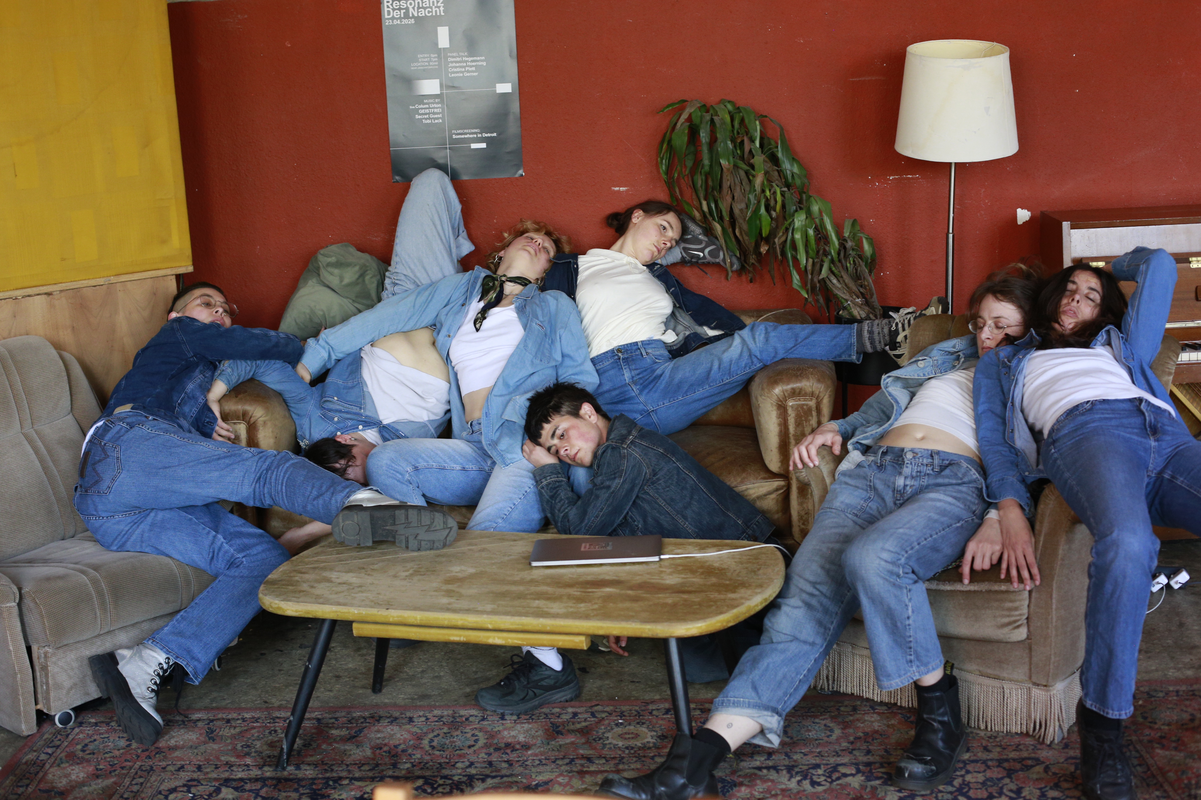 Seven performers in denim piled across a worn sofa, bodies overlapping in a state of collective rest and surrender. Limbs intertwined, faces hidden, the group occupies the couch as a single mass. A low wooden table in the foreground.