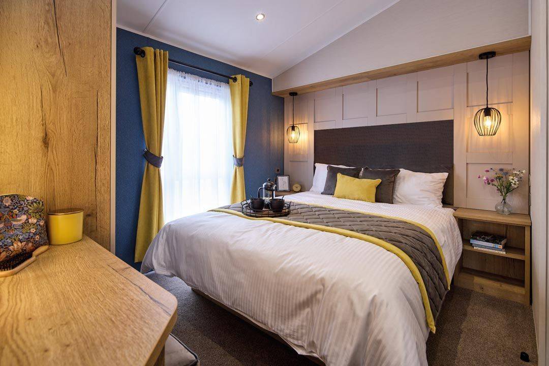 The Willerby Gainsborough master bedroom has two accent walls, a deep blue and a panelled wall, with yellow and grey soft furnishings to match the colour scheme of the home.