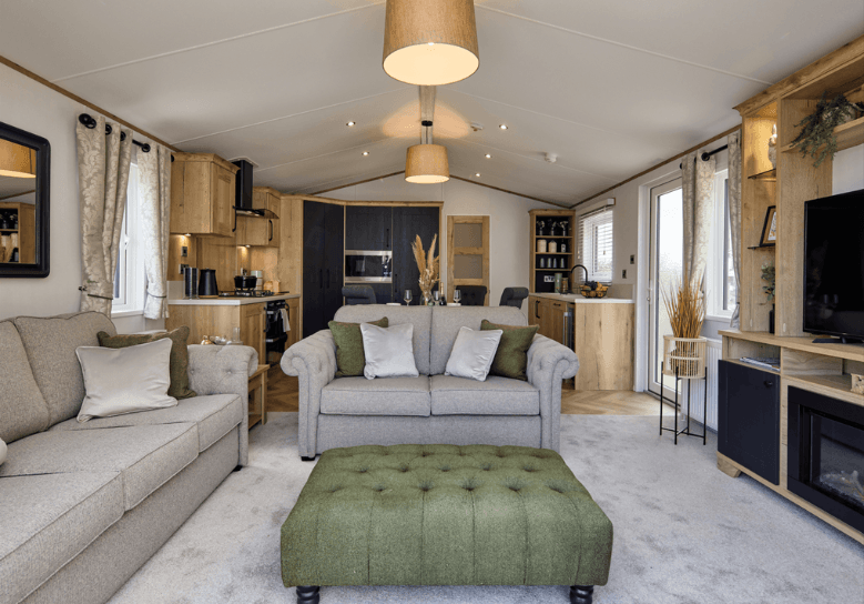 The Willerby Dorchester lounge with grey sofas and a green stylish footstool, with a view of the media unit and kitchen.