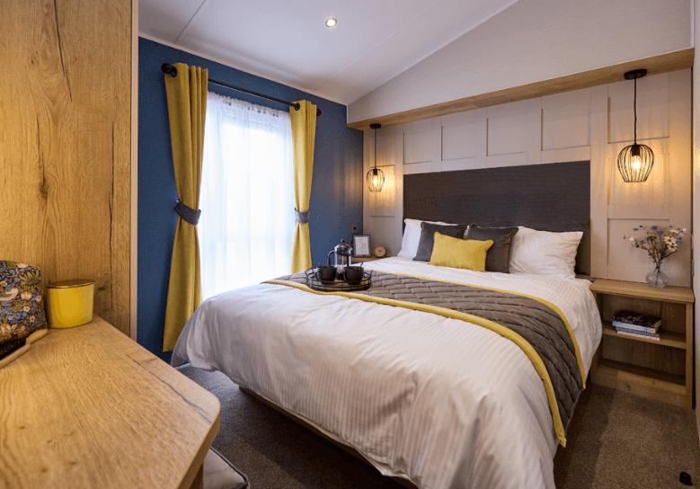 The Willerby Gainsborough master bedroom has two accent walls, a deep blue and a panelled wall, with yellow and grey soft furnishings to match the colour scheme of the home.
