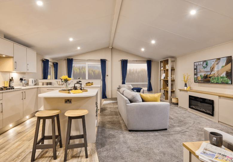 The Willerby Hayward view of the kitchen and lounge, which has a kitchen island in white as the centrepiece with two stools in grey and oak effect.