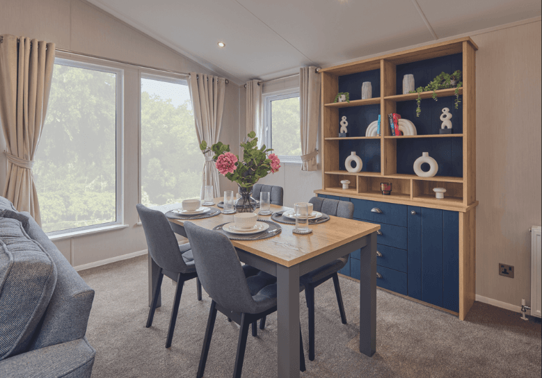 The Willerby New Holland dining area has 4 grey chairs surrounding a grey and oak effect table, with an oak effect and deep blue ornament unit behind.