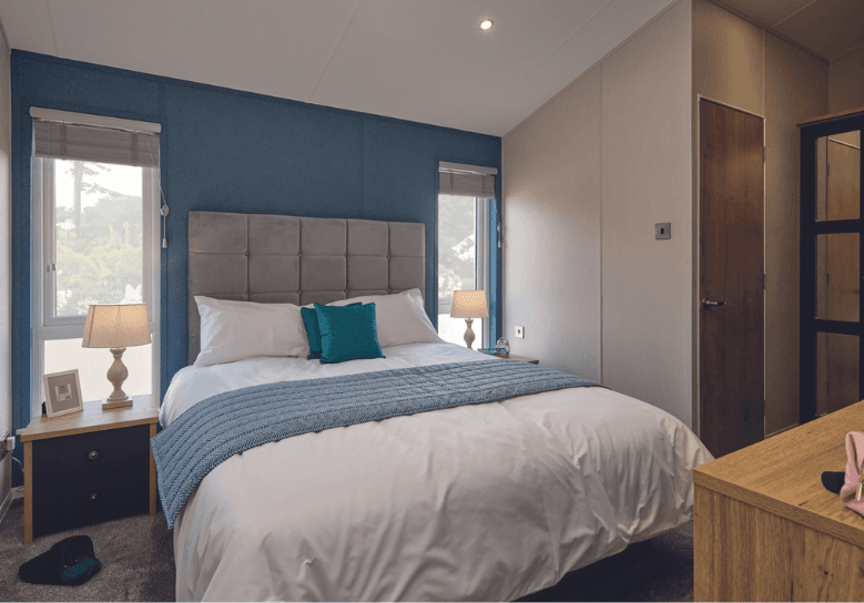 The main bedroom in the Willerby New Holland holiday lodge. The large bed is flanked by two windows and storage units. There is a door leading to the en-suite bathroom. 