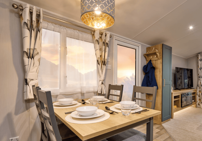 The Willerby Sierra dining area has an oak effect and grey table with grey dining chairs surrounding it, and a patterned gold and grey light shade above.