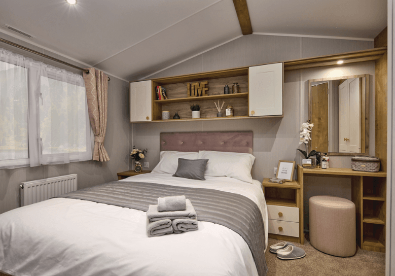 The Willerby Sierra master bedroom has oak effect storage units with white cupboard doors, a matching dressing area and an upholstered pink headboard matching the colour scheme of the home.