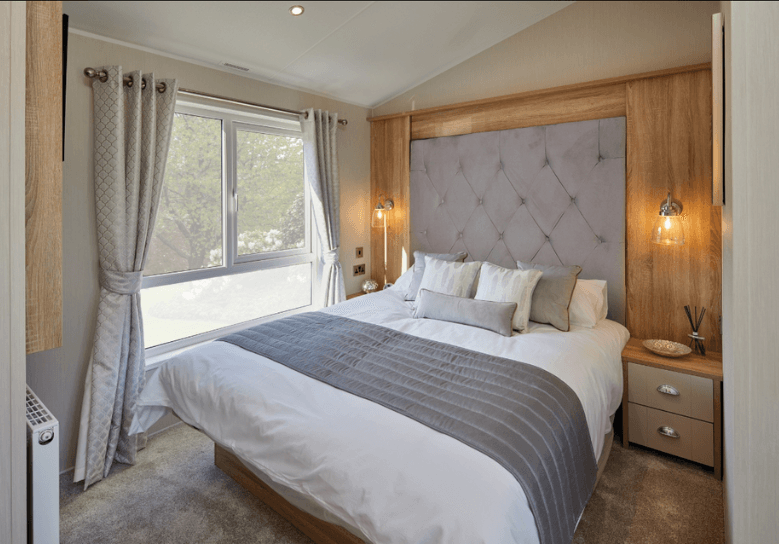 The Willerby Vogue Classique master bedroom has a tall grey upholstered headboard with matching soft furnishings, and a oak effect feature wall.
