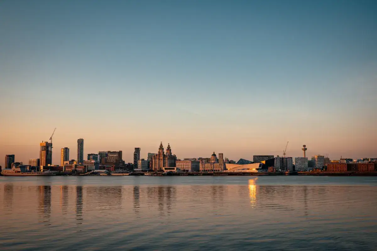 Liverpool skyline from across the Mersey. The sky is deep blue and the low sun casts a golden light on the buildings which are a mixture of modern and classical. 