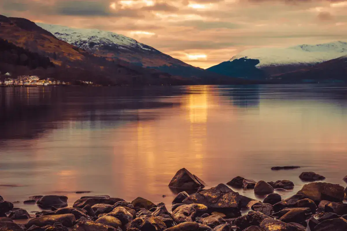 View over Loch Lomond to snow-topped mountains with the sun low in the sky. 