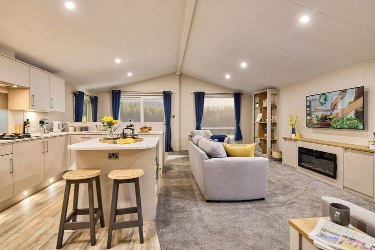 The Willerby Hayward view of the kitchen and lounge, which has a kitchen island in white as the centrepiece with two stools in grey and oak effect.