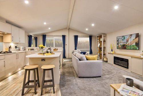 The Willerby Hayward view of the kitchen and lounge, which has a kitchen island in white as the centrepiece with two stools in grey and oak effect.