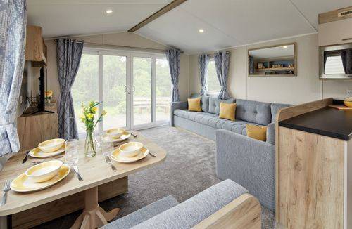 The Willerby Linwood lounge has a grey L-shaped fixed sofa with yellow scatter cushions, and a view of the media unit and dining table.